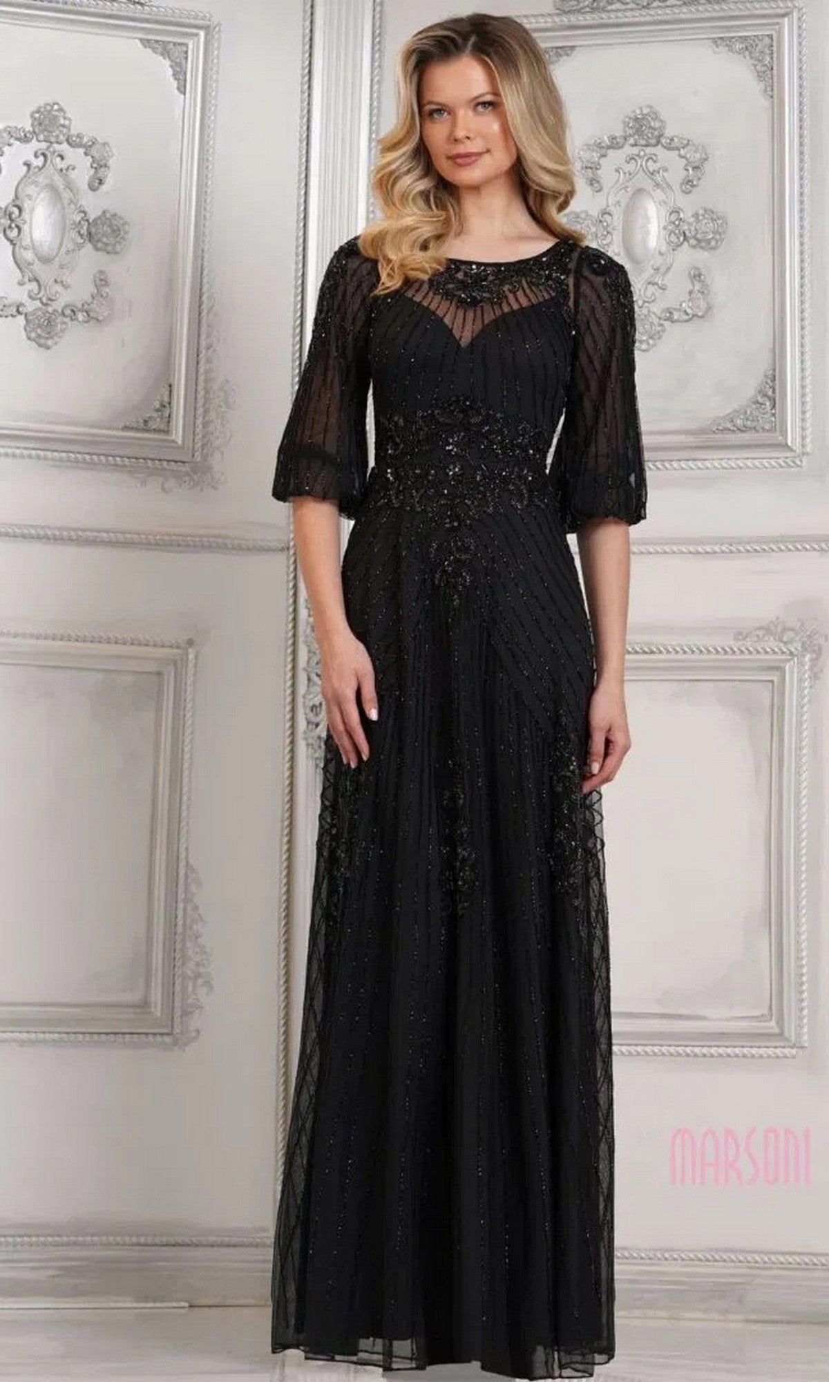 A woman in a black Long Formal Dress: Marsoni MV1323 with sheer sleeves and intricate beadwork stands against an ornate white wall.